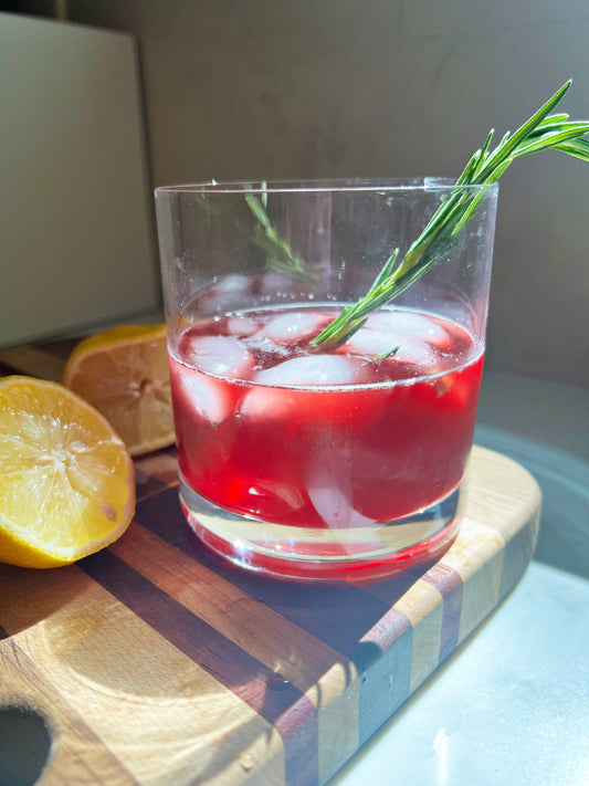 Glass with red cherry mocktail and rosemary sprig on a wooden cutting board with a cut lemon