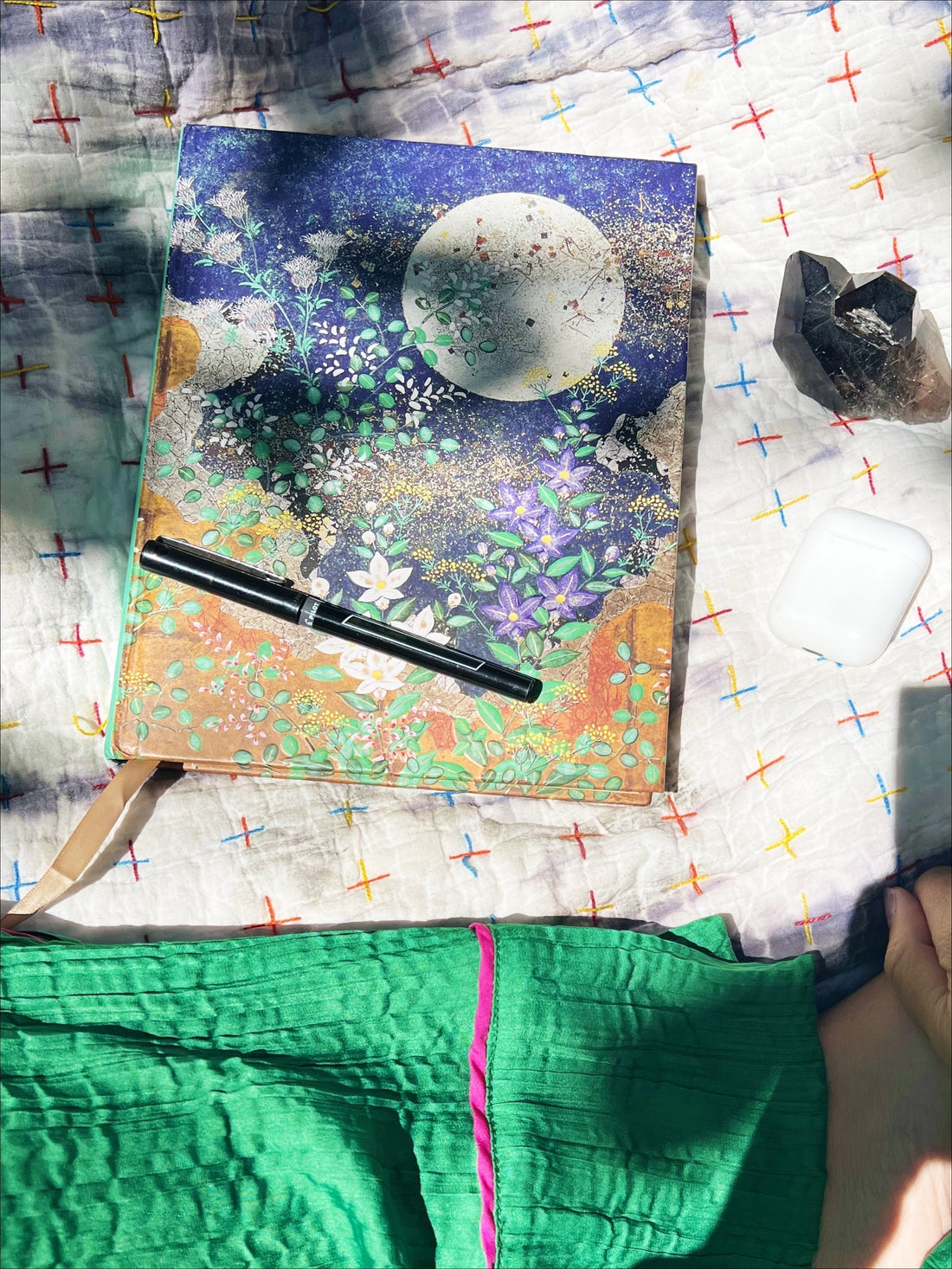 Journal with black pen on blanket with smoky quartz, headphones and green silk pants for moment to reset and relax the nervous system