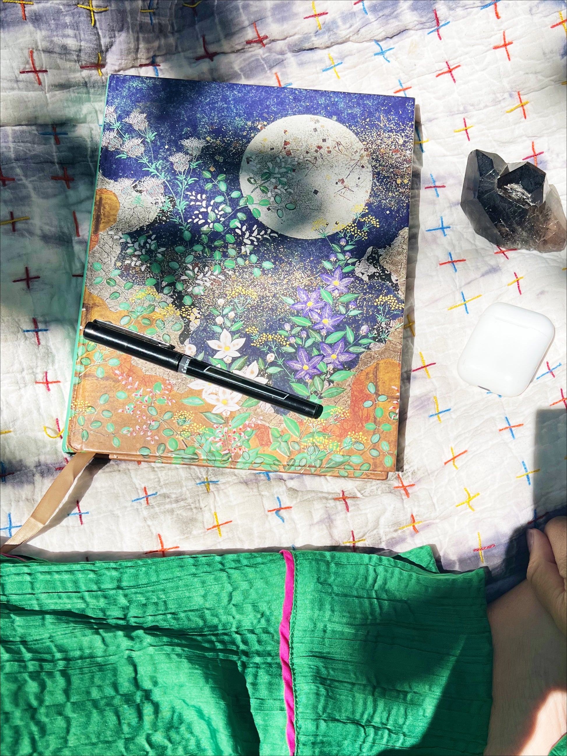 Journal with moon and greens on blanket with tree shadow next to black pen and smoky quartz crystal and green silk pants