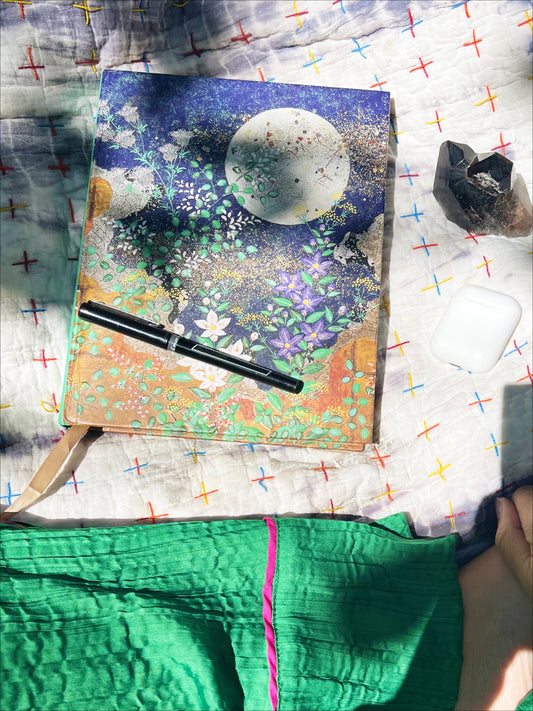 Journal with moon and greens on blanket with tree shadow next to black pen and smoky quartz crystal and green silk pants
