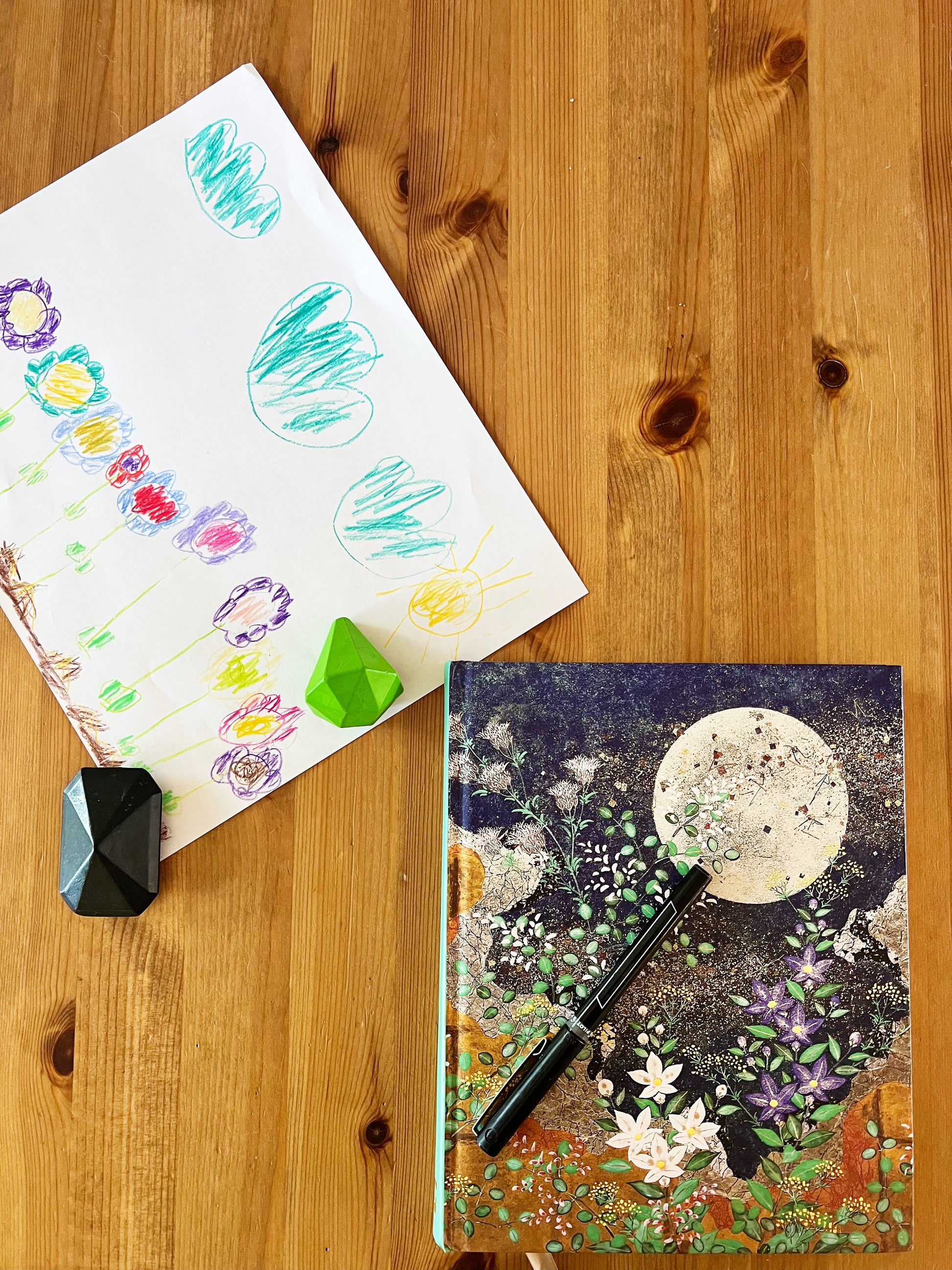 Journal with moon and greens with black pen on table next to child’s artwork and crystal crayons